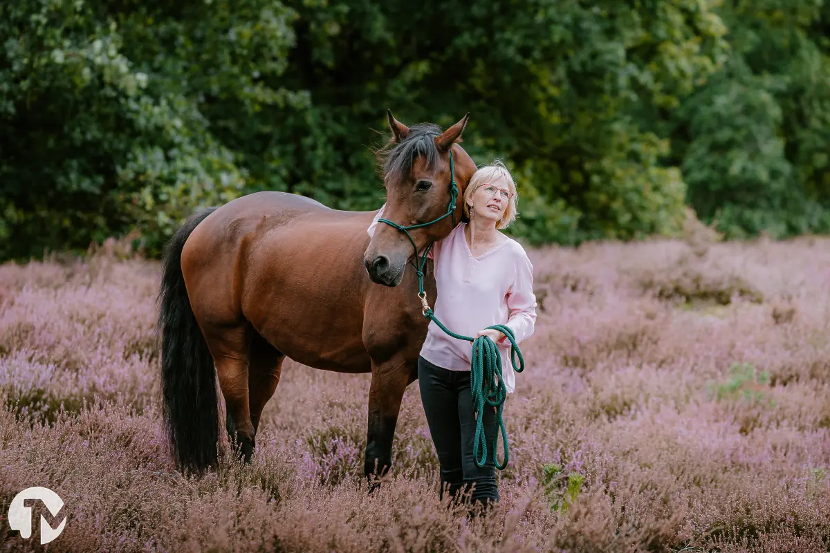 vrouw houdt paard bij hoofd vast en kijkt dromerig weg in de heide